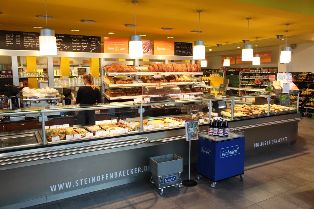 Mitarbeiterin an der Backshop-Theke des Bioladens, dahinter Brotregale, rechts die Obstabteilung